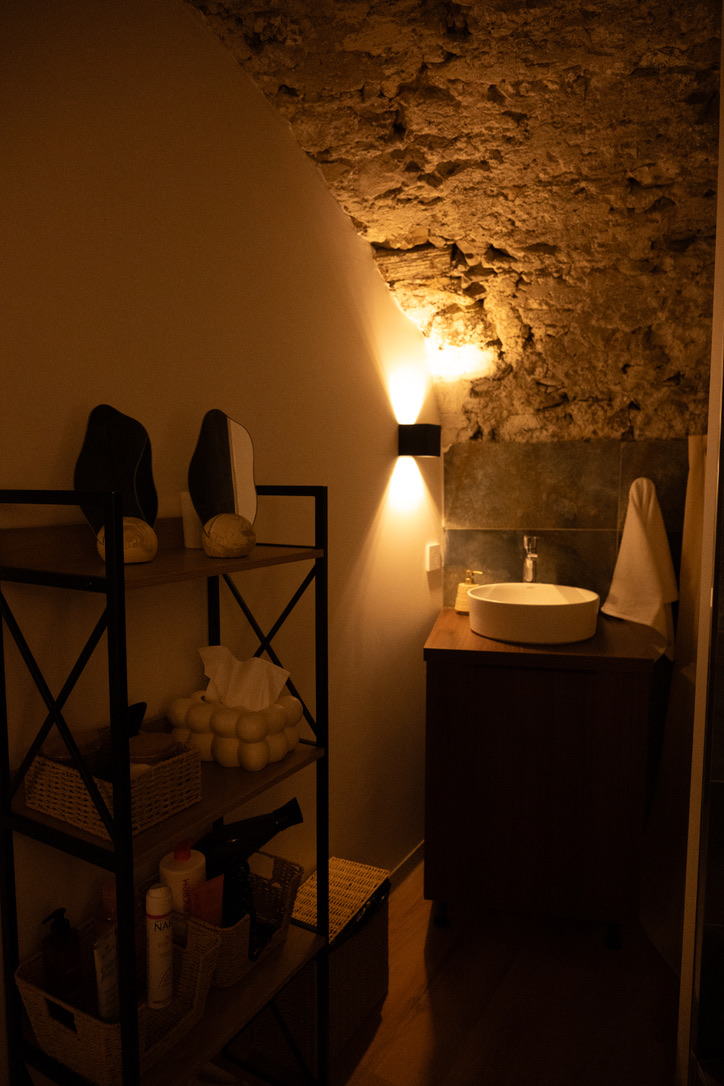 Coin salle d'eau du studio LOUMA sous voûte en pierre apparente, applique allumée et vasque